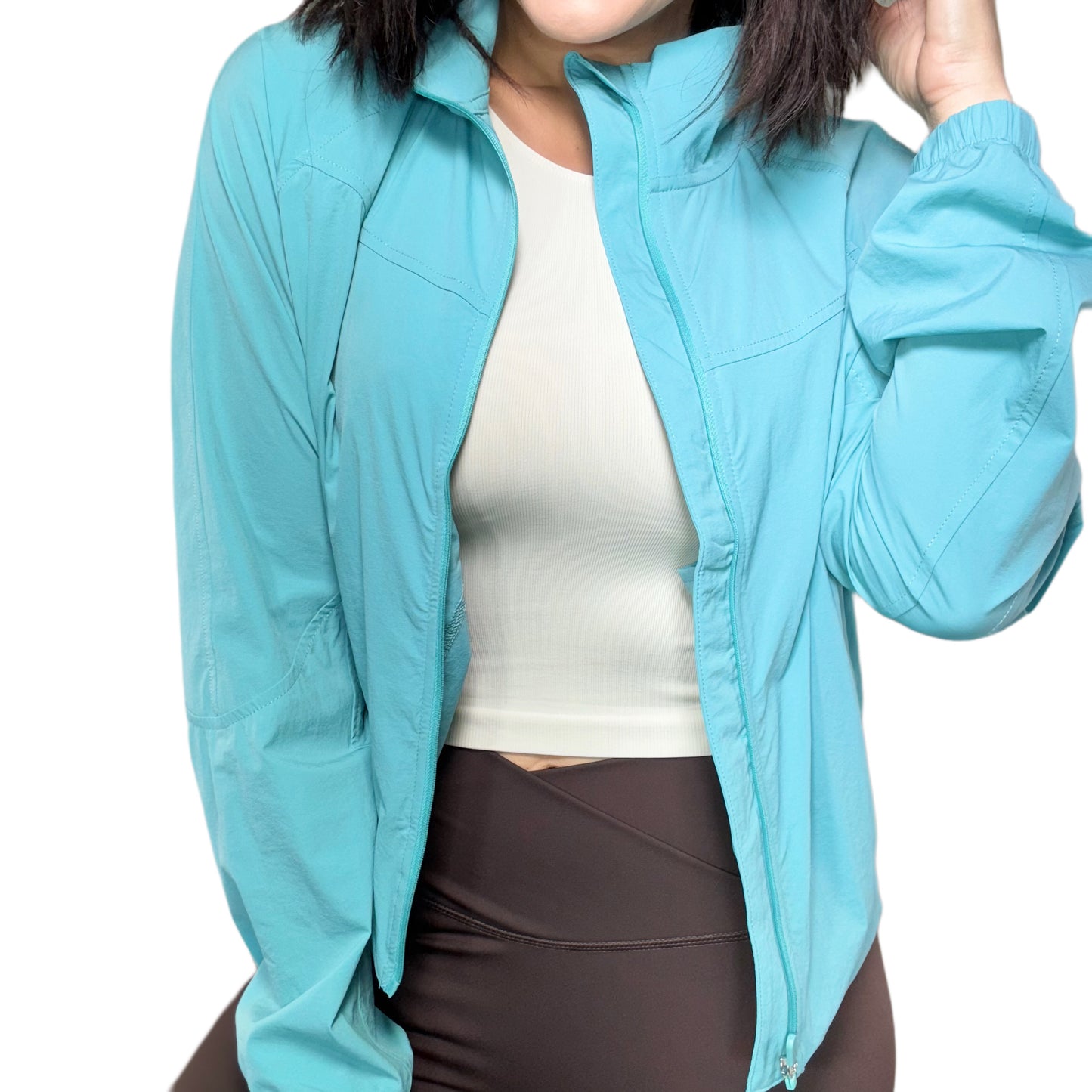 Woman wearing a aqua athletic jacket, open over a tan shirt and brown athletic pants on a white background