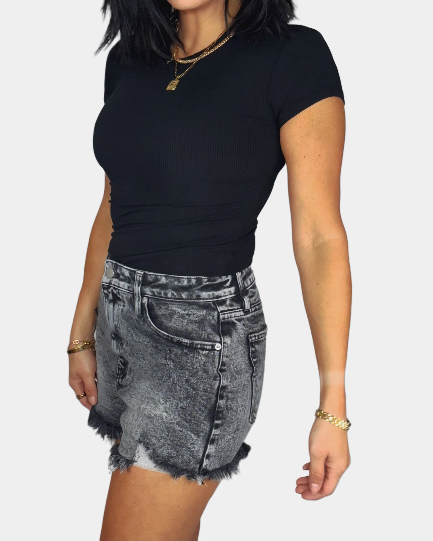 Side profile of a woman wearing a black ribbed bodysuit and distressed denim shorts on a white background