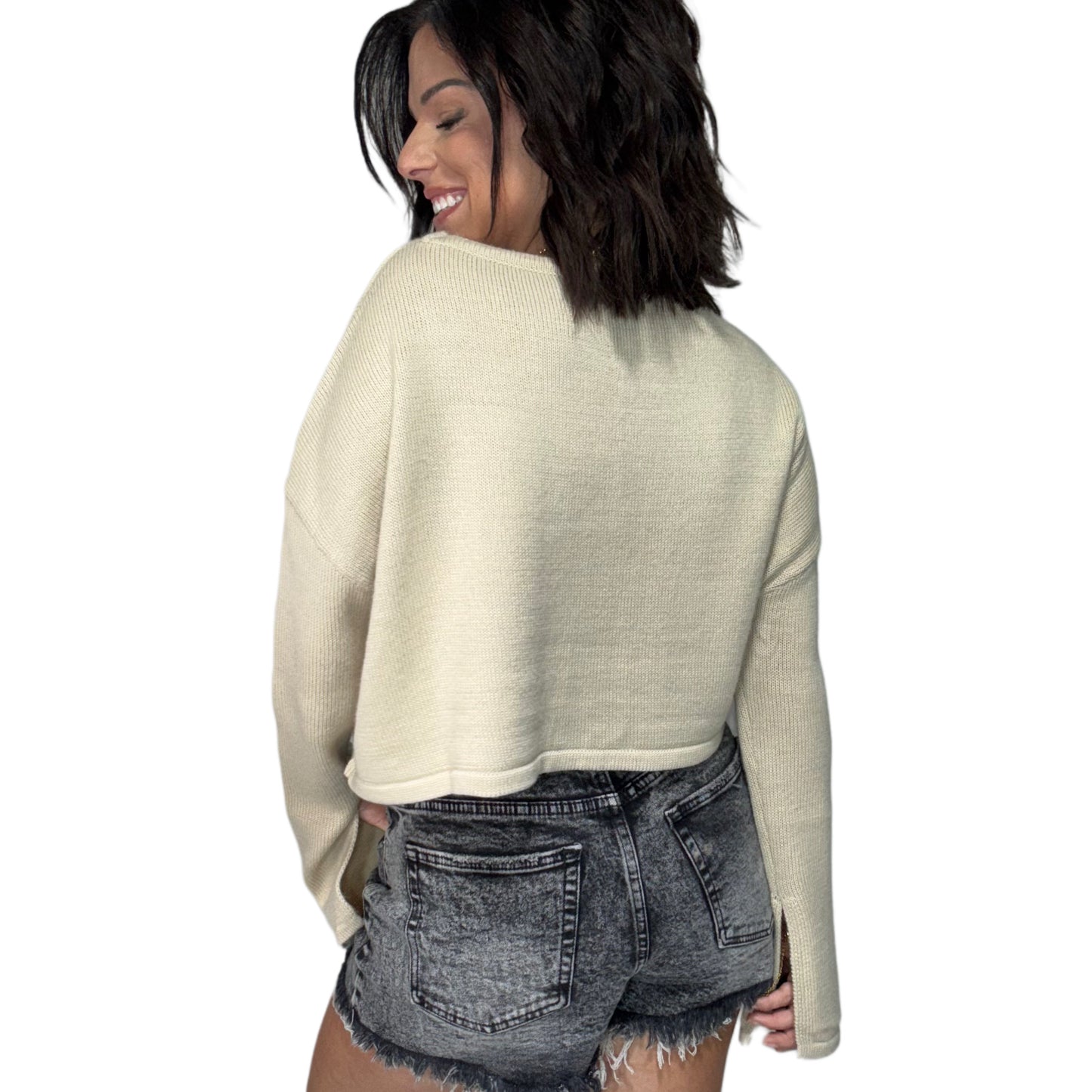 Woman turned around wearing a tan long sleeve knit top and black denim shorts on a white background