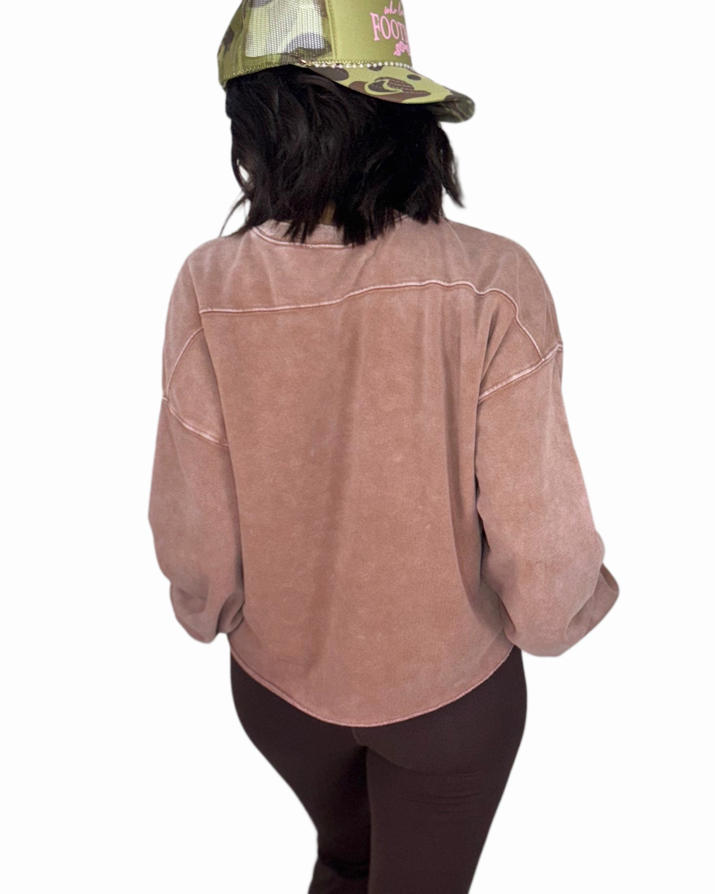 Woman turned around wearing a mauve sweatshirt and brown leggings with a green cap on a white background