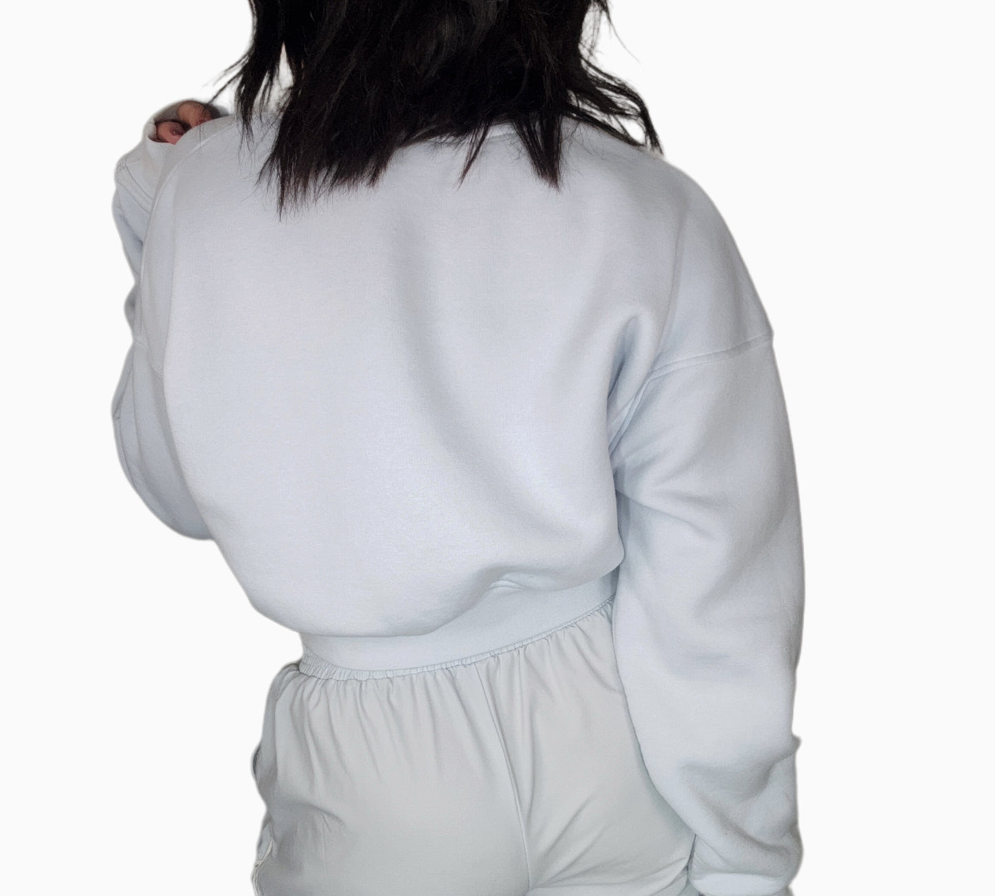 Back profile of a woman wearing a light blue cropped sweatshirt with matching track pants, on a white background