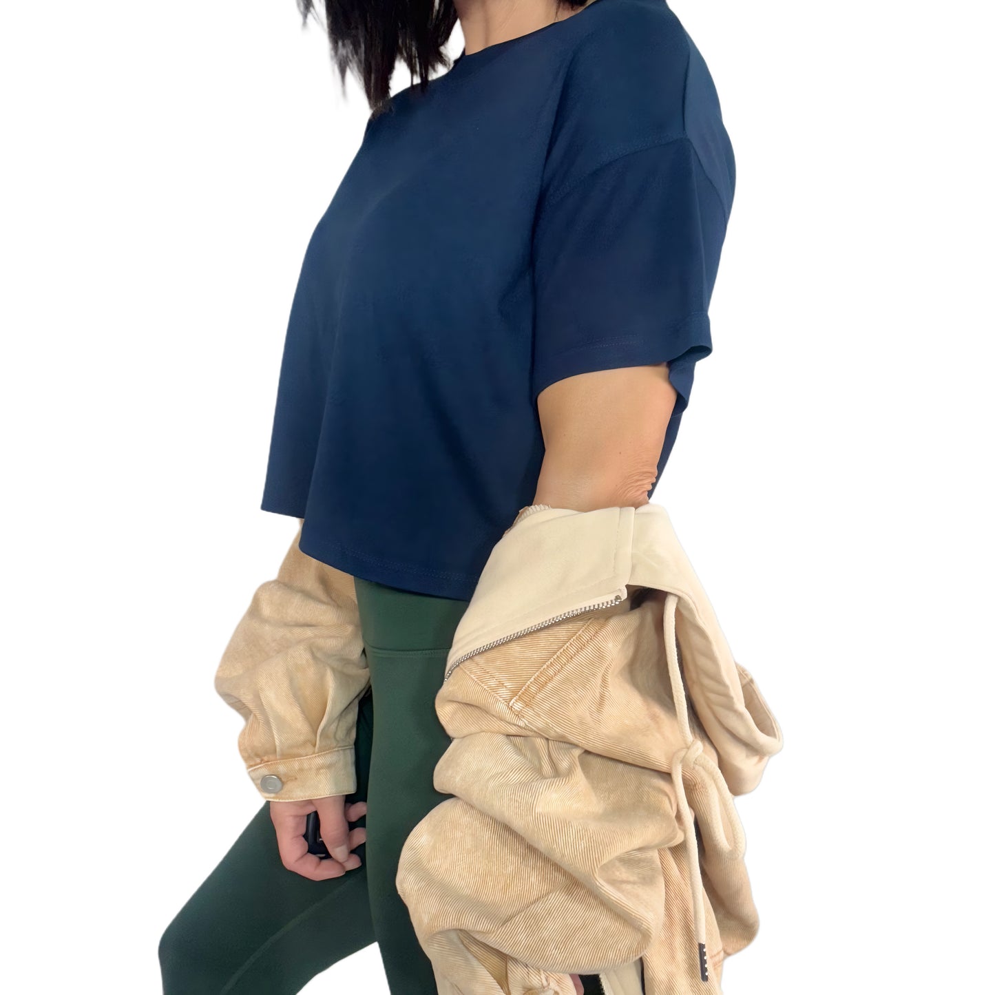 Side profile of a woman wearing a navy cropped tee and green active pants with beige jean jacket on a white background