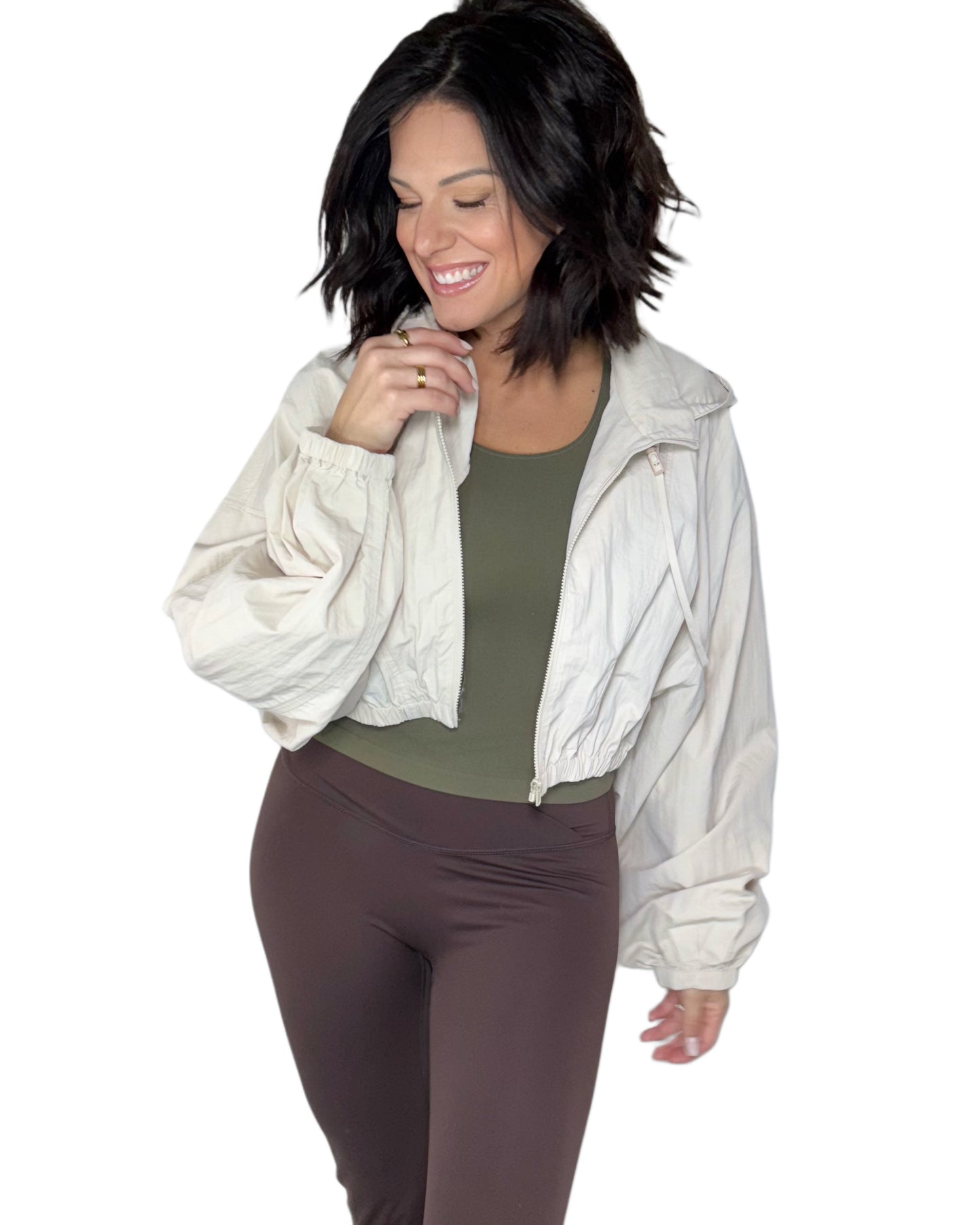 Woman wearing a tan windbreaker jacket over a green top and brown active pants on a white background