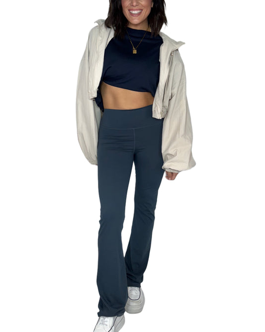 Woman wearing navy bootcut leggings with a dark navy top and tan jacket on a white background