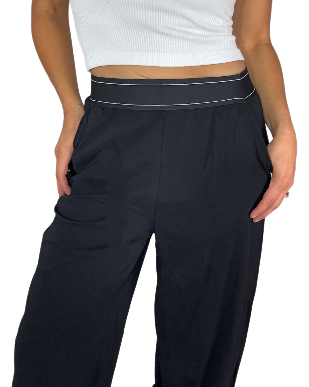 Close up of a woman modeling a pair of navy blue wide-leg sweatpants with a thin white stripe detail along the waistband, paired with a white crop top and white sneakers, against a plain white background.