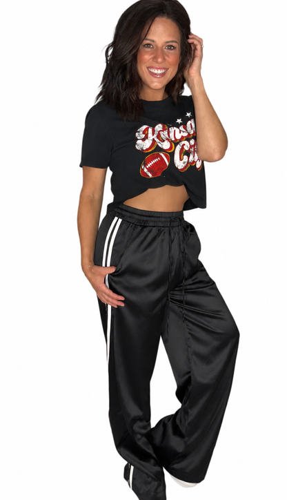 A woman modeling a black cropped t-shirt with a football graphic and matching black satin track pants with white side stripes, set against a plain white background.