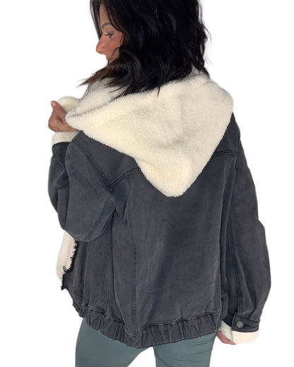 A woman wearing a dark gray washed denim jacket with a thick cream sherpa fleece hood lining, shown from a back-three-quarter angle against a plain white background.
