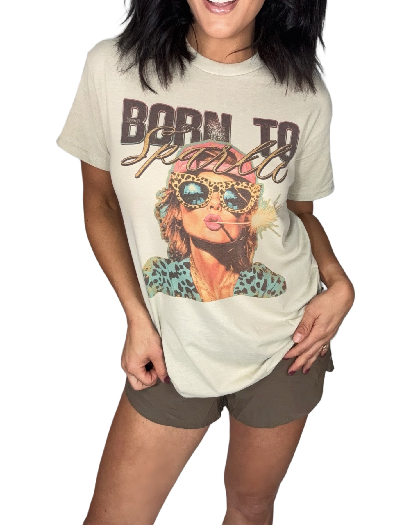 Woman wearing a sand color t-shirt with a "Born to Sparkle" graphic on the front, brown shorts and a white background 