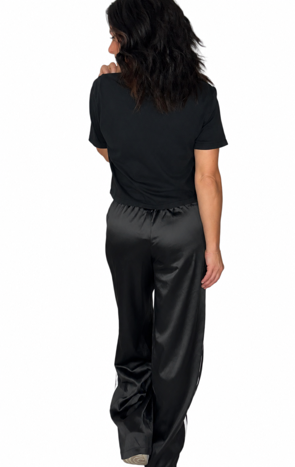 A person shown from the back wearing a black short-sleeve top and shiny black satin or silky pants against a plain white studio background.