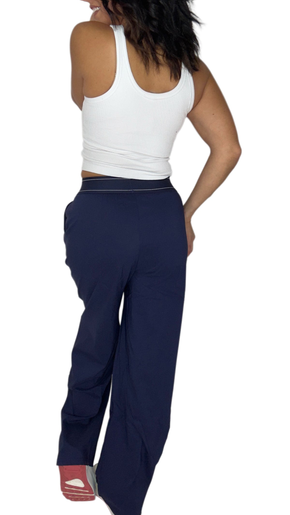 Back profile of a woman modeling a pair of navy blue wide-leg sweatpants with a thin white stripe detail along the waistband, paired with a white crop top and white sneakers, against a plain white background.