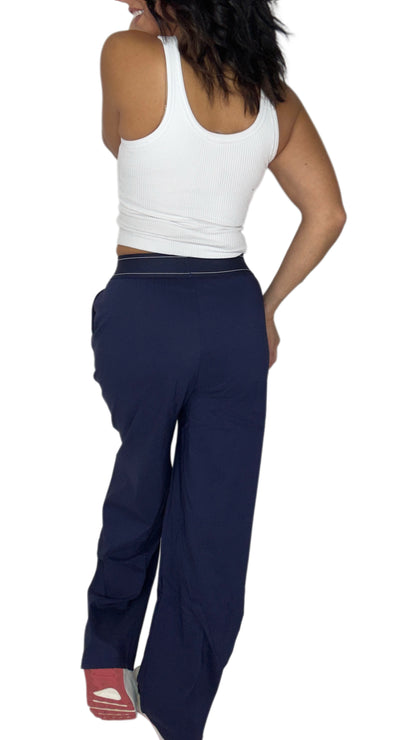 Back profile of a woman modeling a pair of navy blue wide-leg sweatpants with a thin white stripe detail along the waistband, paired with a white crop top and white sneakers, against a plain white background.
