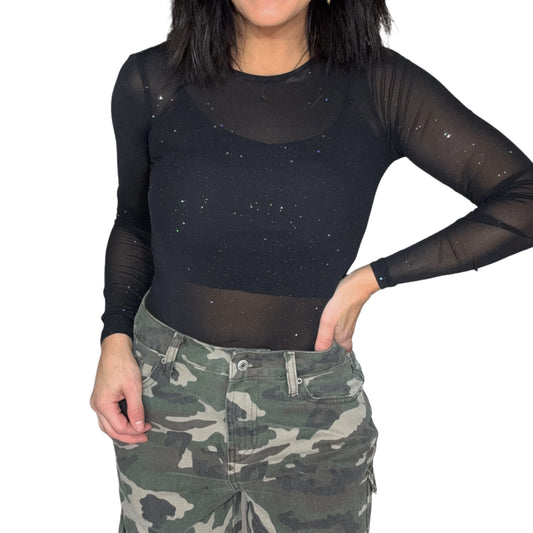 Close up of a woman wearing a black mesh long-sleeve top with silver specks on it and camouflage pants on a white background