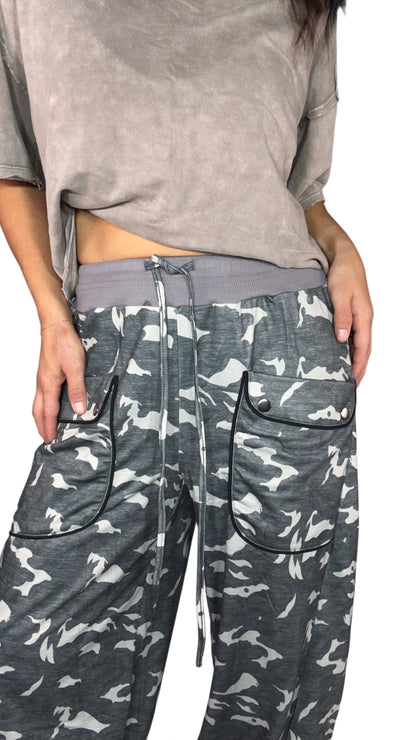Close up of a woman wearing relaxed lounge camouflage carog pants with a thick grey waist band and front pockets with a brown tee, on a white background. 