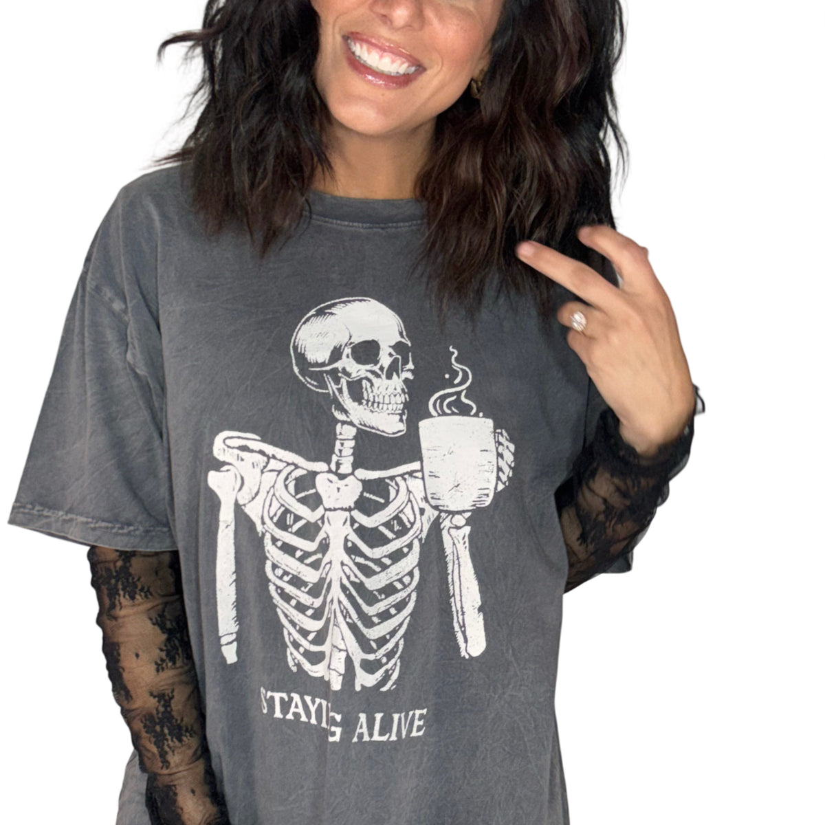 Close up of a woman wearing a deep grey Halloween skeleton tee that says "staying alive" with a lace undershirt on white background 