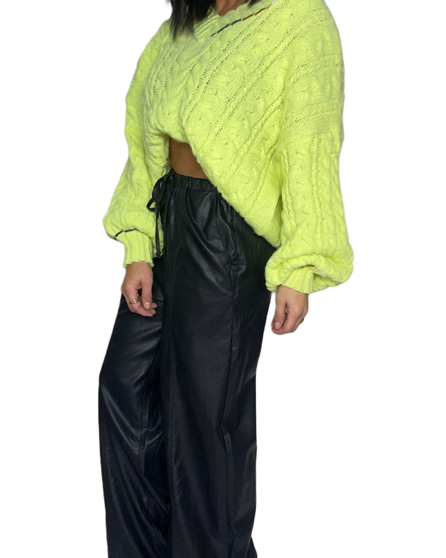 Side profile of a woman wearing a bright neon cable knit sweater and black pants on a white background