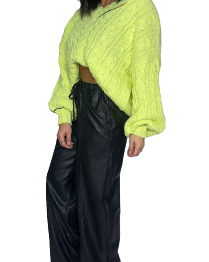 Side profile of a woman wearing a bright neon cable knit sweater and black pants on a white background