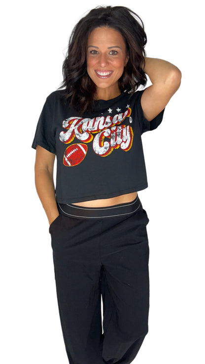 A woman modeling a black short-sleeve crop top with a colorful 'Kansas City' graphic and football motif, paired with black sweatpants against a plain white background.