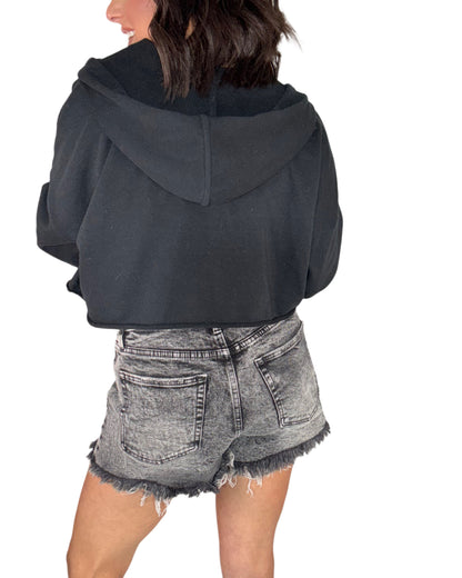 Woman wearing a black cropped raw hem hoodie and denim shorts on a white background
