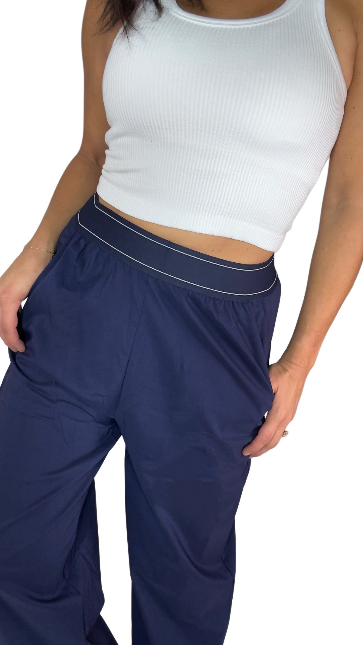 Close up of a woman  modeling a pair of navy blue wide-leg sweatpants with a thin white stripe detail along the waistband, paired with a white crop top and white sneakers, against a plain white background.