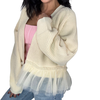 Side profile of a woman wearing a cream cardigan with tulle at the bottom over a pink top and jeans on a white background
