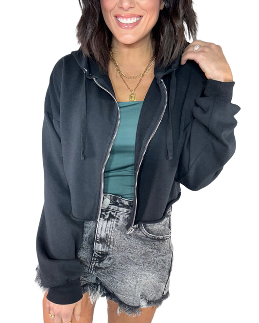 Woman wearing a black raw hem zip-up hoodie over a teal top and denim shorts on a white background