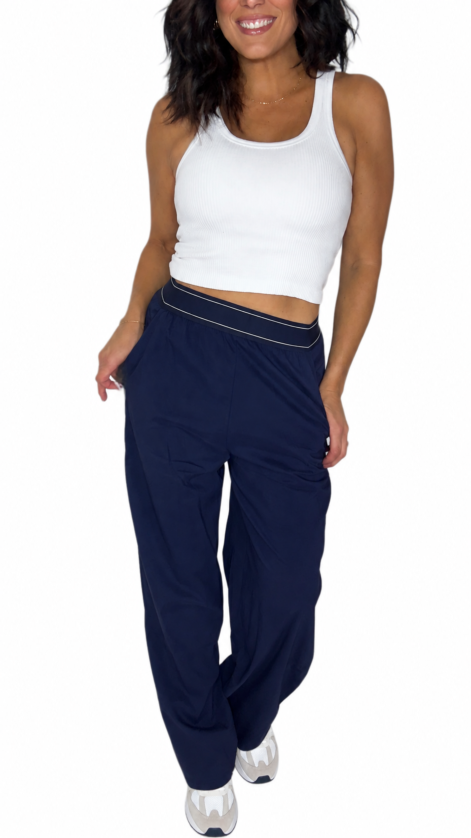 A woman modeling a pair of navy blue wide-leg sweatpants with a thin white stripe detail along the waistband, paired with a white crop top and white sneakers, against a plain white background.