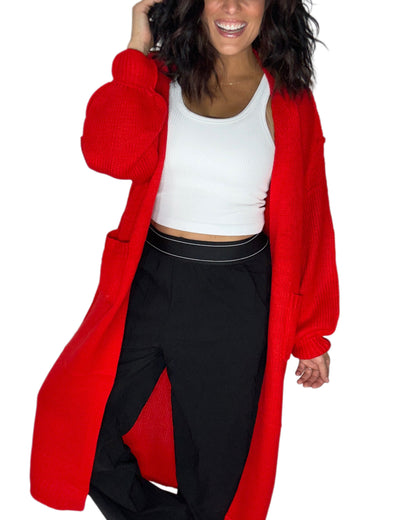 A woman wearing a bright red open-front knit cardigan over a white crop top and black pants, shown against a plain white background.