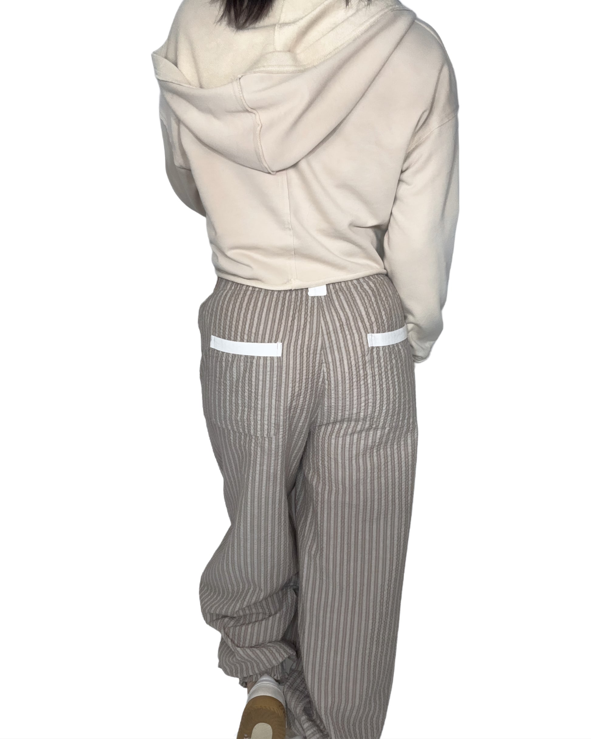 Back profile of a woman wearing a beige cropped hoodie and striped pants on a white background