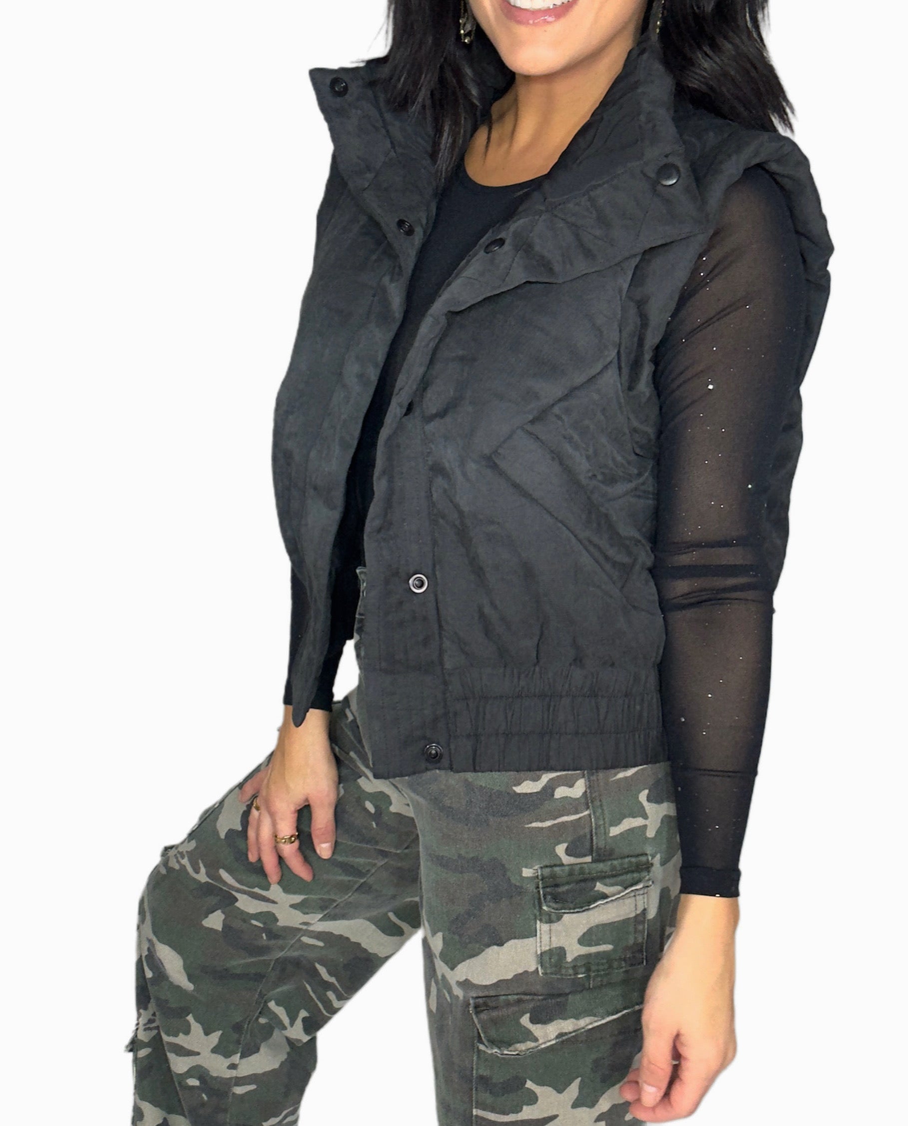 Side profile of a woman wearing a black vest over a black shirt with camouflage pants on a white background