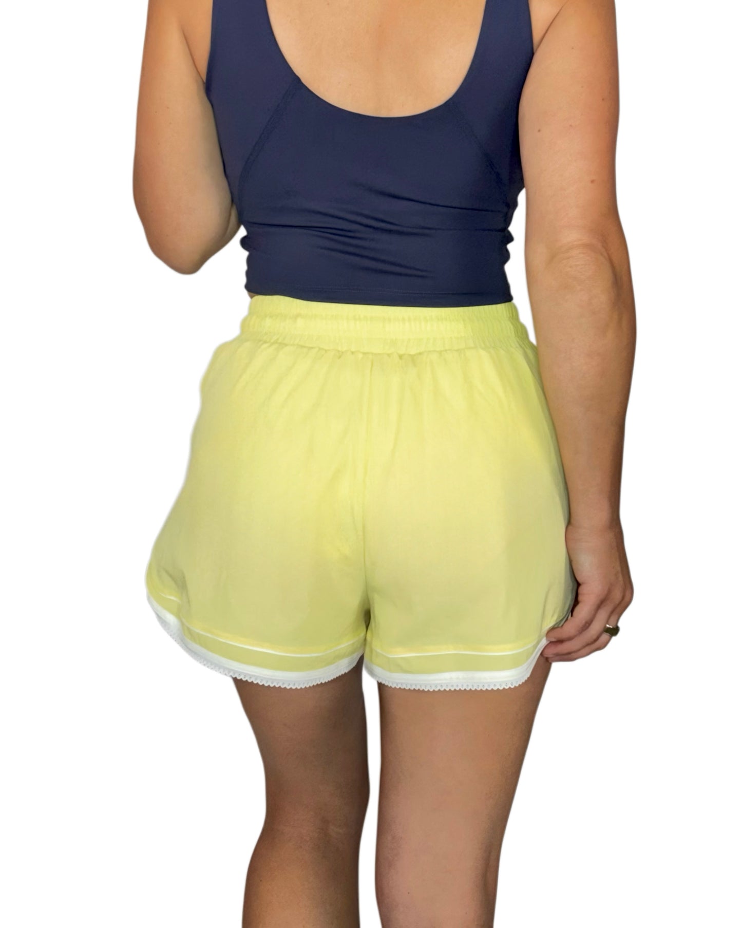 Back profile of a woman wearing pale yellow active shorts with a navy tank top, on a white background 