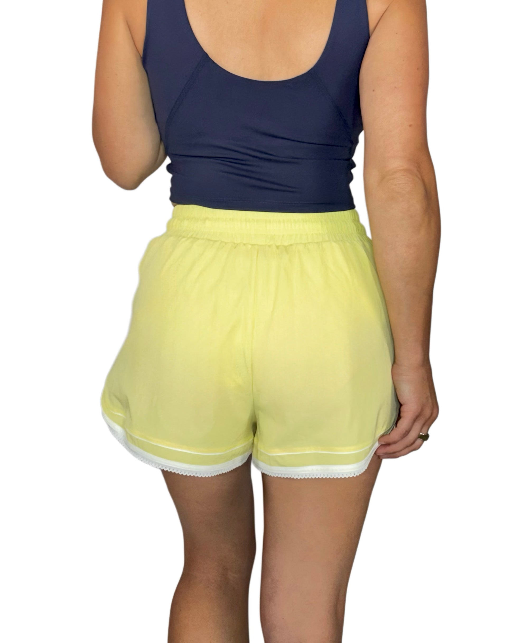 Back profile of a woman wearing pale yellow active shorts with a navy tank top, on a white background 