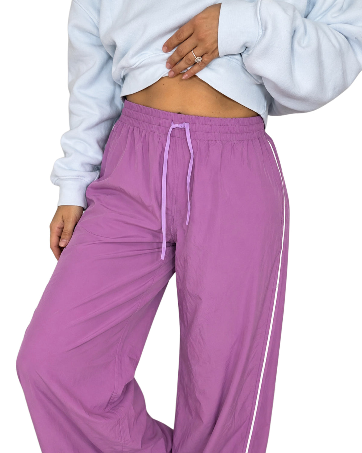 Close up of a woman wearing mulberry color track pants with a drawstring and white line going down the side with a light blue cropped sweatshirt on a white background