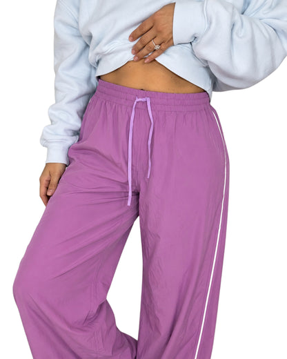 Close up of a woman wearing mulberry color track pants with a drawstring and white line going down the side with a light blue cropped sweatshirt on a white background