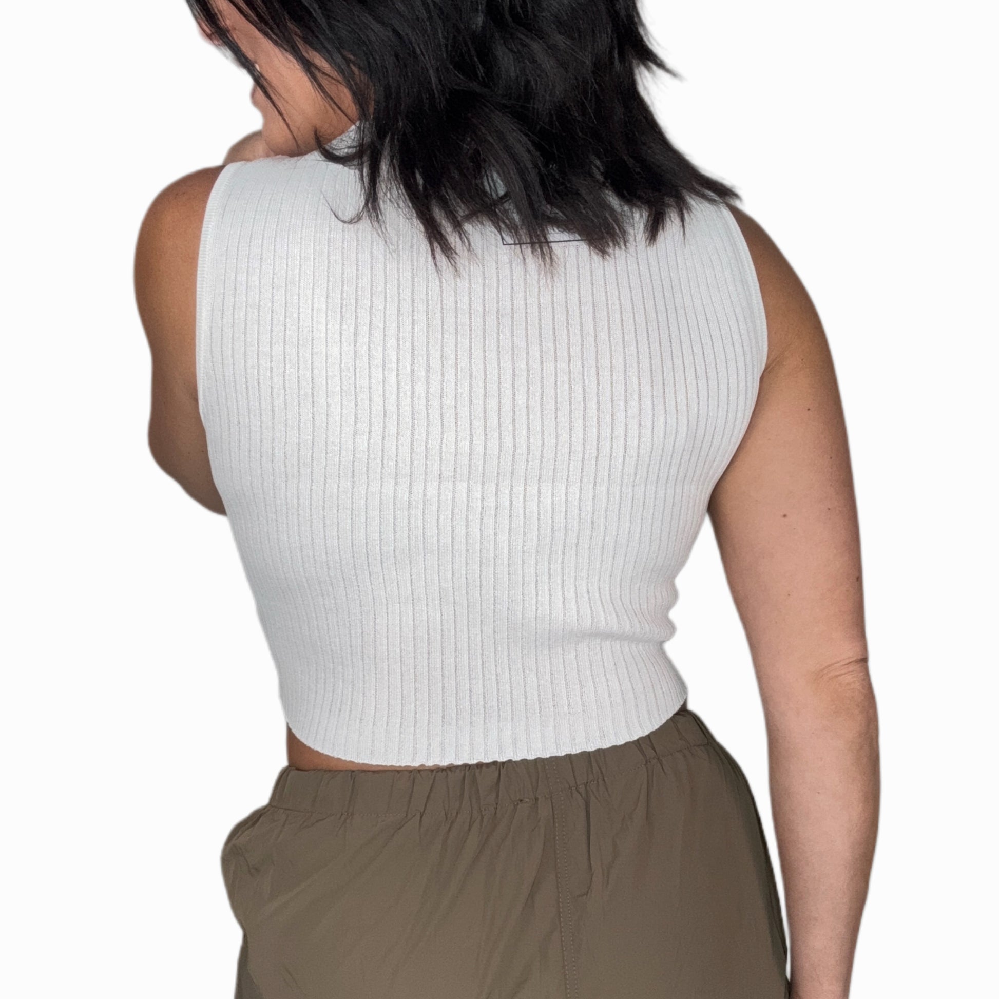 Back profile of a woman wearing a white knit tank top with green pants and a white background 