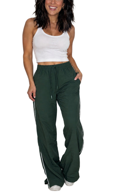 Woman wearing dar green track pants with a drawstring and a white line detailing down the side with a ribbed white tank top, on a white background 