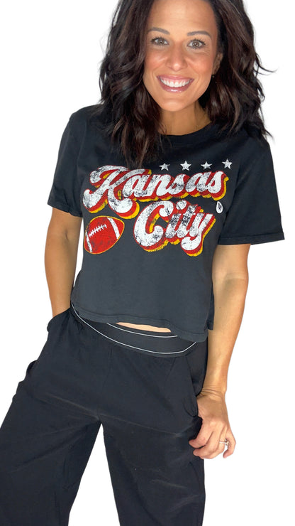 A woman modeling a black short-sleeve crop top with a distressed 'Kansas City' graphic in white, red, and yellow, paired with black pants against a plain white background.