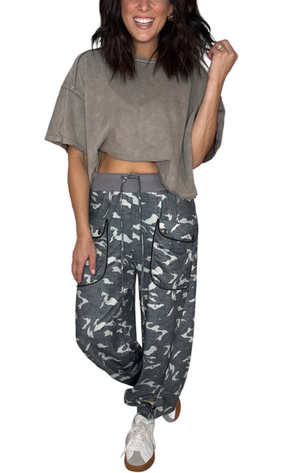 Woman wearing  relaxed lounge camouflage cargo pants with a thick grey waist band and front pockets with a brown tee, on a white background. 