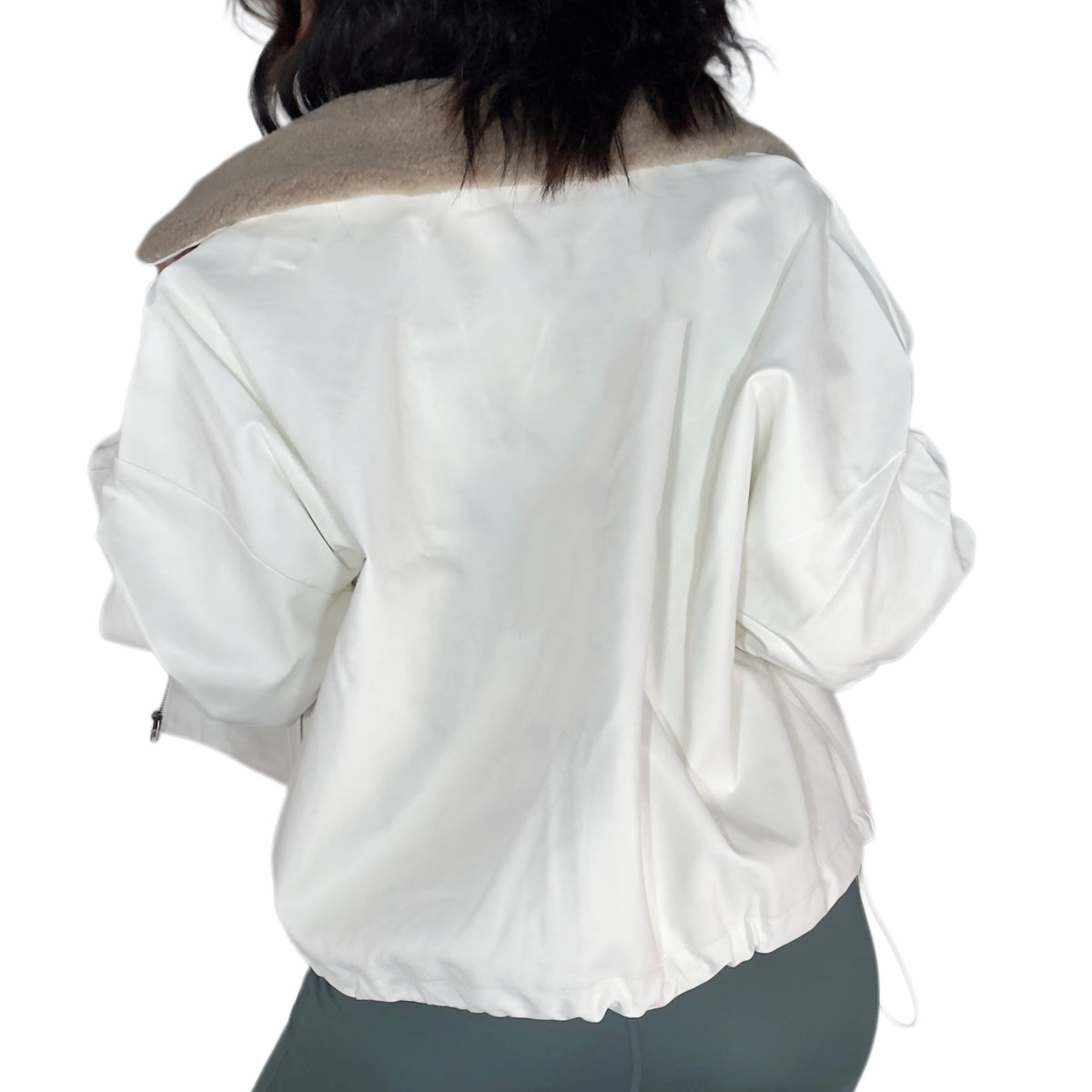 Back profile of a woman wearing an off white jacket with tan sherpa around the collar with green active pants, on a white background 