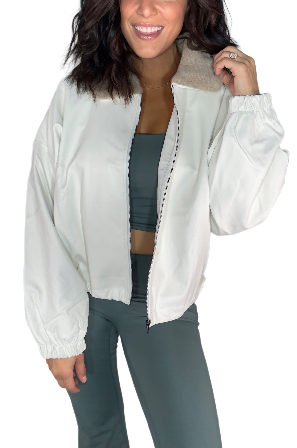 A woman wearing a white lightweight zip-up jacket with a soft beige sherpa collar, paired with a dark gray sports bra and gray leggings, against a plain white background.