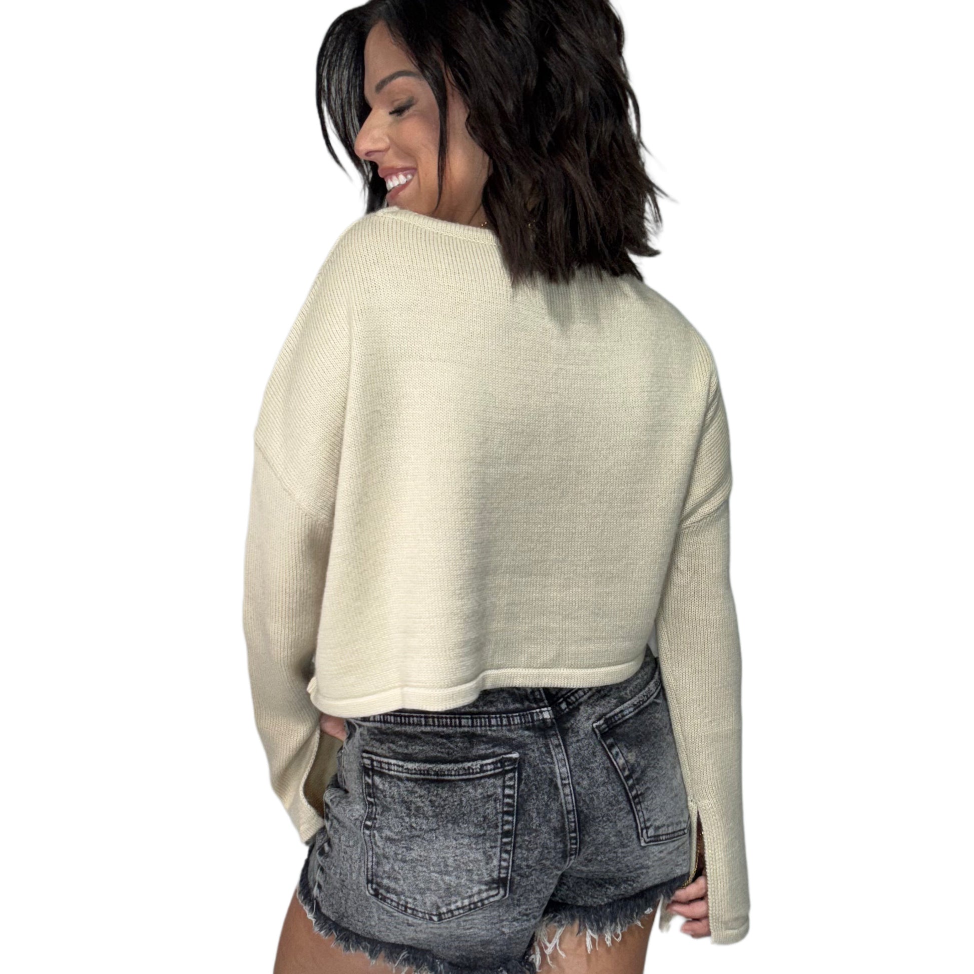 Woman turned around wearing a tan long sleeve knit top and black denim shorts on a white background