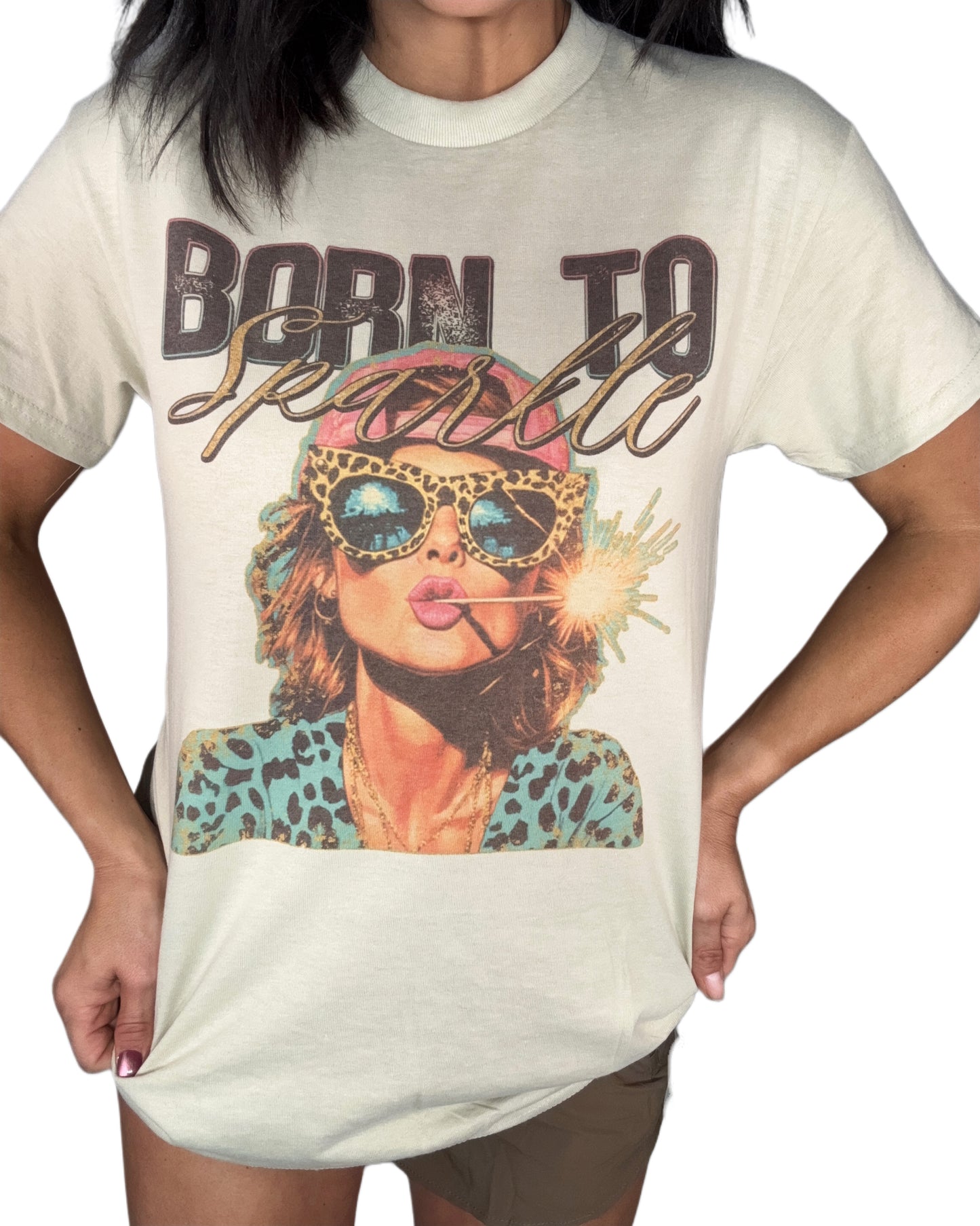 Close up of a woman wearing a sand color t-shirt with a "Born to Sparkle" graphic on the front, brown shorts and a white background 