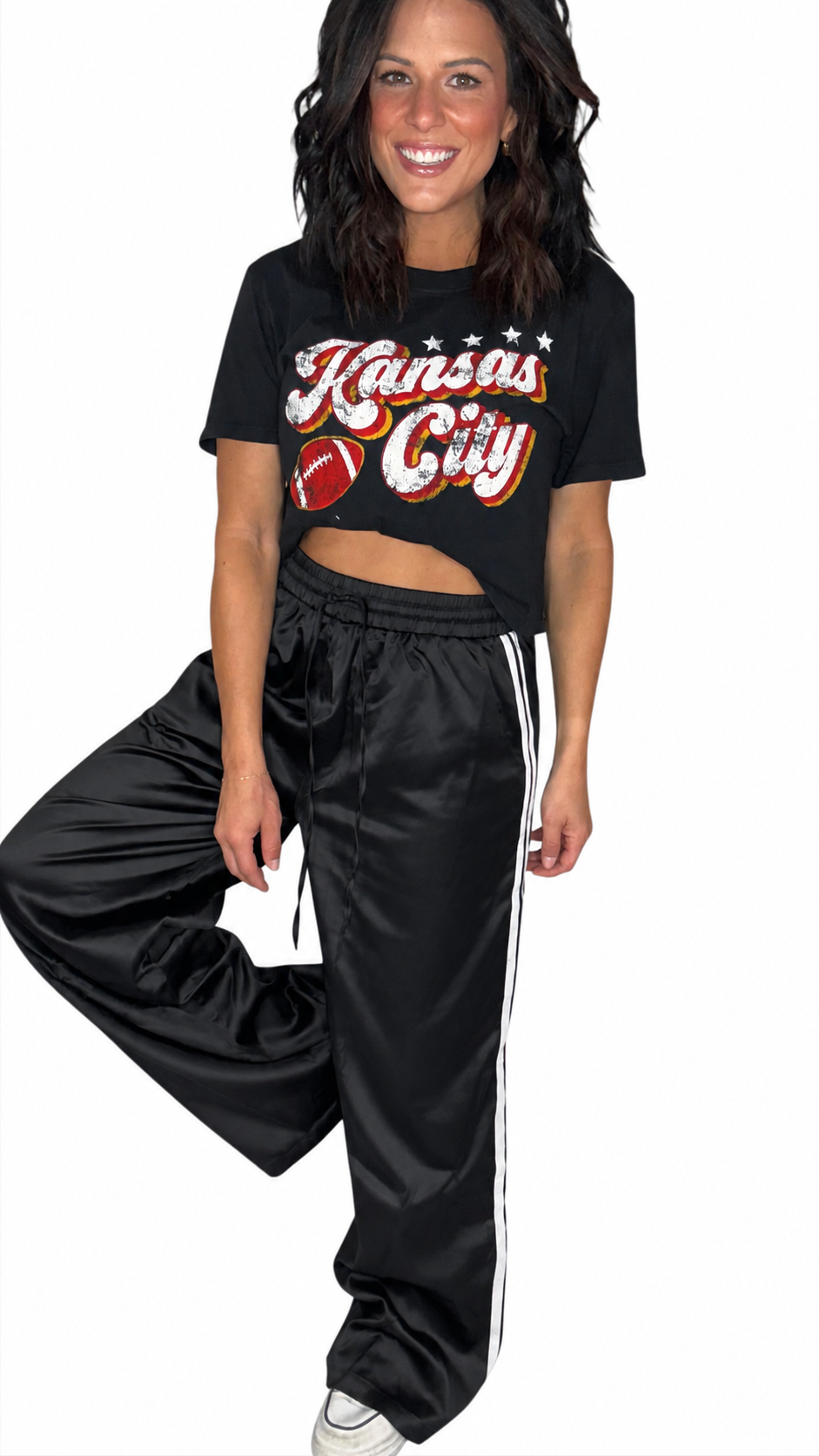 A woman modeling a black cropped graphic T-shirt and black satin track pants with white side stripes. The shirt features 'Kansas City' in white script with red and orange outlining and a football graphic.