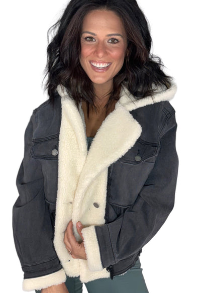 A woman modeling a dark gray washed denim jacket with a thick cream sherpa fleece lining, paired with olive green pants, against a solid white background.