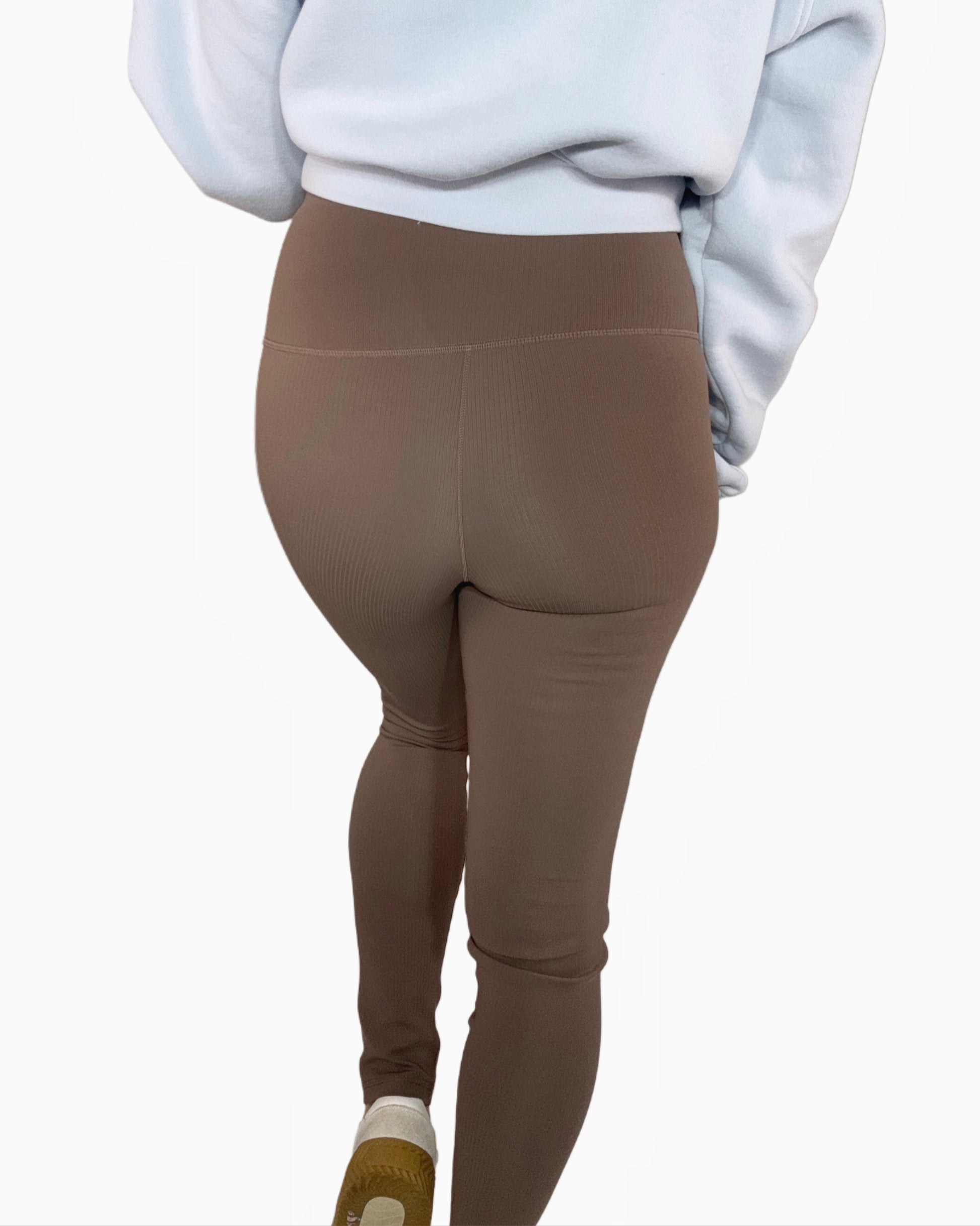 Back profile of a woman wearing ribbed deep taupe leggings, a cropped light blue sweatshirt, on a white background.