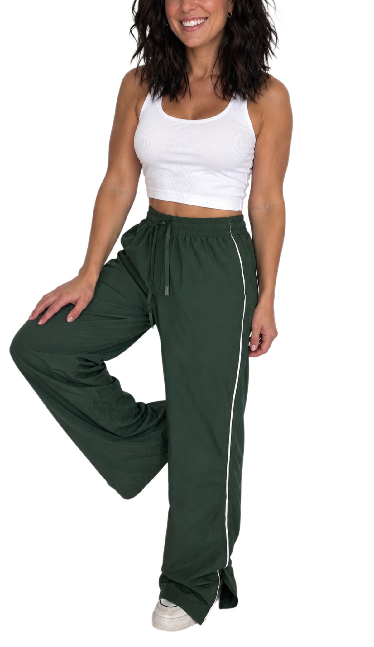Side profile of a woman wearing dar green track pants with a drawstring and a white line detailing down the side with a ribbed white tank top, on a white background 