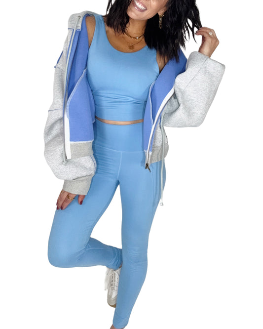 Woman wearing a powder blue tank top and leggings set with grey and blue sweat jacket, on a white background 