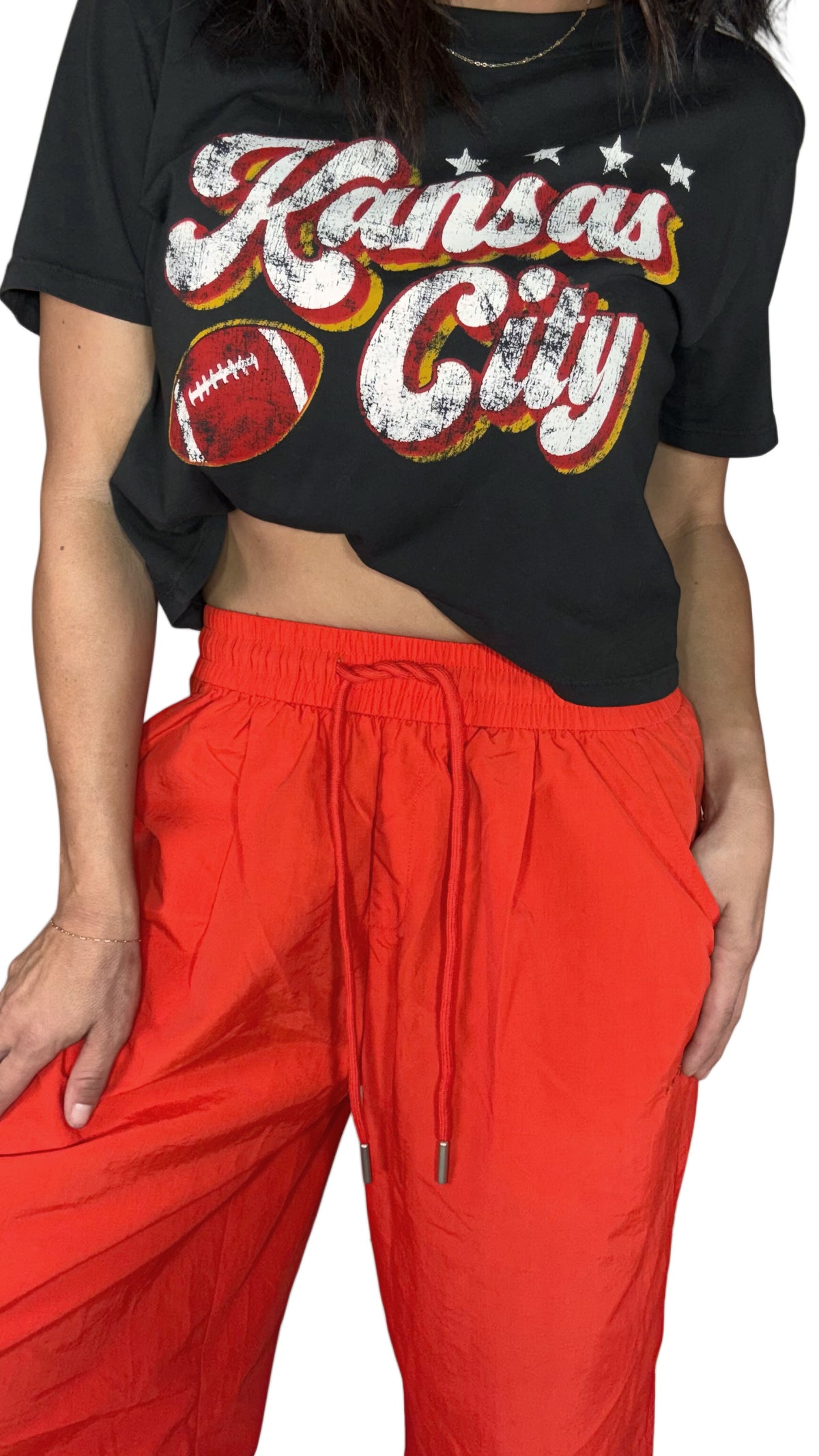 A cropped view of a person wearing a black graphic T-shirt with distressed white 'Kansas City' lettering, a football graphic, and red/orange/yellow shadowing, paired with bright red drawstring pants against a plain white background.