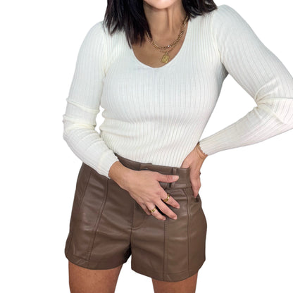 Close up of a woman wearing a cream v neck top and brown leather shorts on a white background
