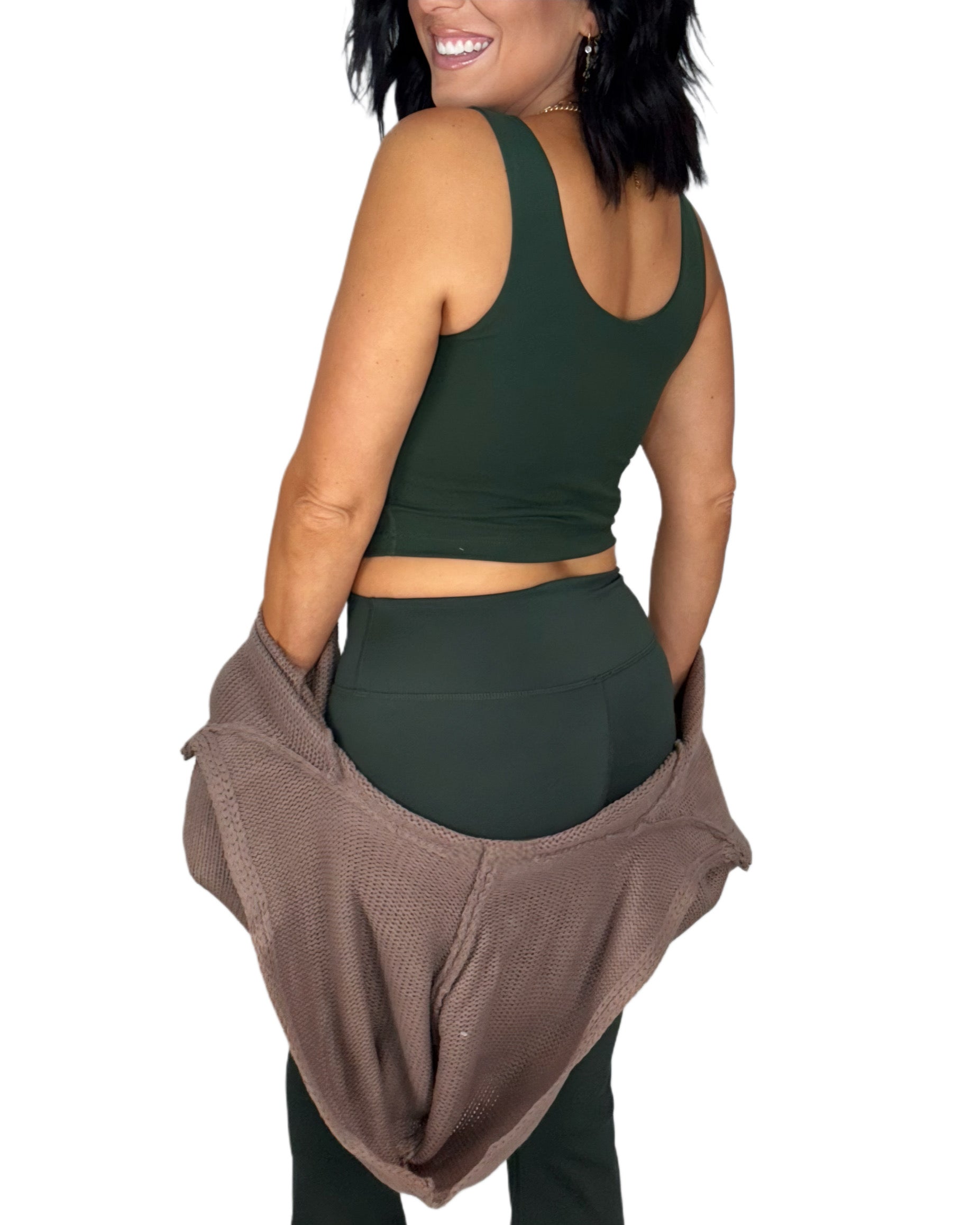 Woman turned around wearing a green athletic outfit with a brown knit jacket  on a white background