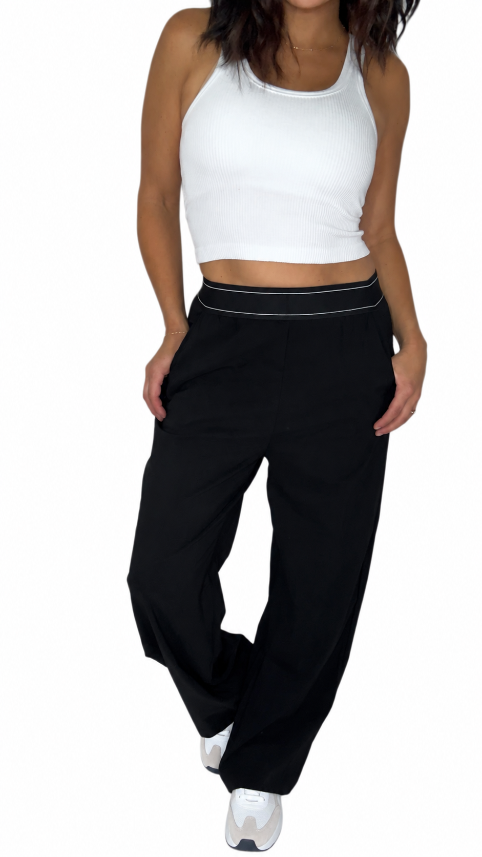 A woman modeling a pair of black wide-leg sweatpants with a thin white stripe detail along the waistband, paired with a white crop top and white sneakers, against a plain white background.